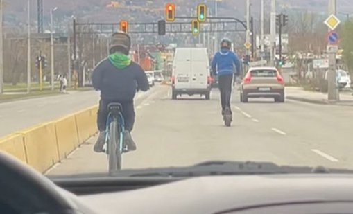 Две деца на тротинетка и колело препускат през трафика на оживен булевард в София 