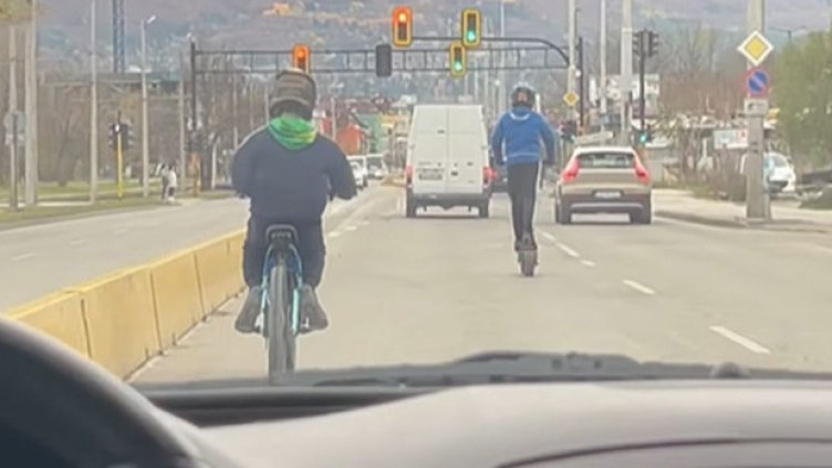 Две деца на тротинетка и колело препускат през трафика на оживен булевард в София 