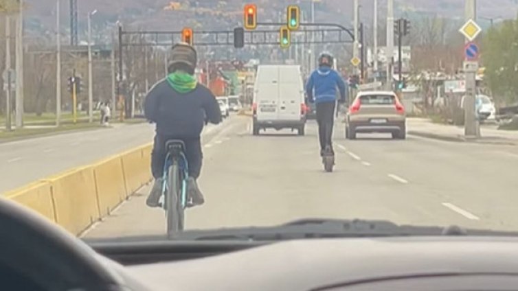 Две деца на тротинетка и колело препускат през трафика на оживен булевард в София 