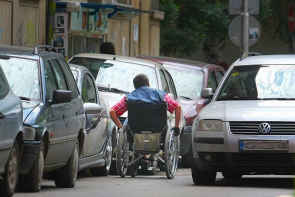 Човек в инвалидна количка се опитва да мине на централна софийска улица днес, 9 юни. Понеже тротоарът е зает от паркирани коли, се налага той да се движи по пътното платно. Снимка: БГНЕС